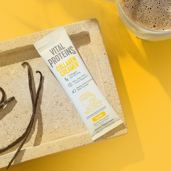 Single serving packet of Collagen Creamer - Vanilla | Vital Proteins placed on a textured tray next to vanilla beans, with a cup of coffee on a yellow surface, highlighting wellness supplements.