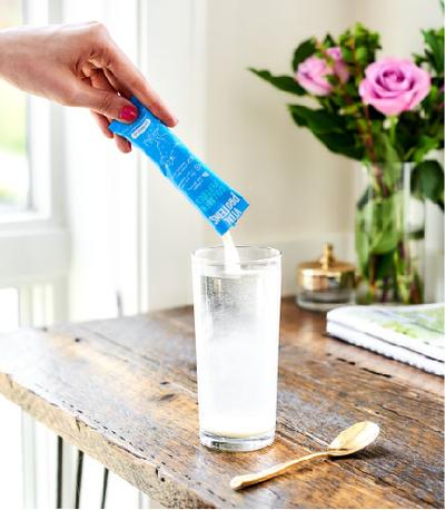 A hand pouring Collagen Peptides - Unflavored | Vital Proteins powder from a blue packet into a clear glass of water on a wooden table with flowers and a golden spoon nearby.