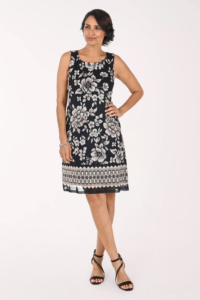 A woman modeling a sleeveless black and white floral print dress with a geometric patterned hem, paired with black strappy heels, against a plain white studio background.
