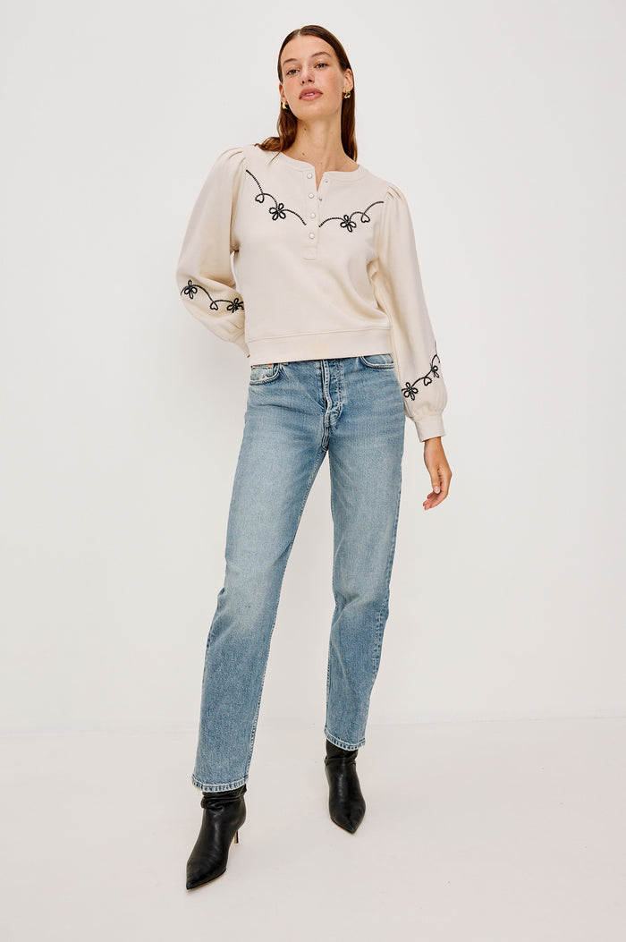 The Hanne is a henley style long sleeve sweatshirt with a snap front placket, rib trim, western style contrast black embroidery and puff sleeves in Ecru colorway. Front full body view featured on model.