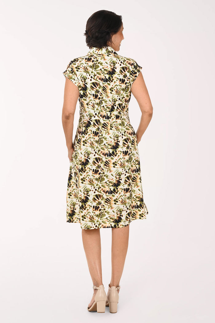 A woman modeling a knee-length floral dress with a cream base and green, black, and brown botanical print, paired with beige heeled sandals against a plain white studio background.
