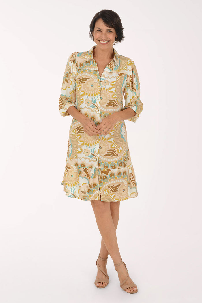 A woman modeling a short-sleeve button-up dress with a beige, gold, and light blue paisley pattern, paired with beige heeled sandals, against a plain white studio background.