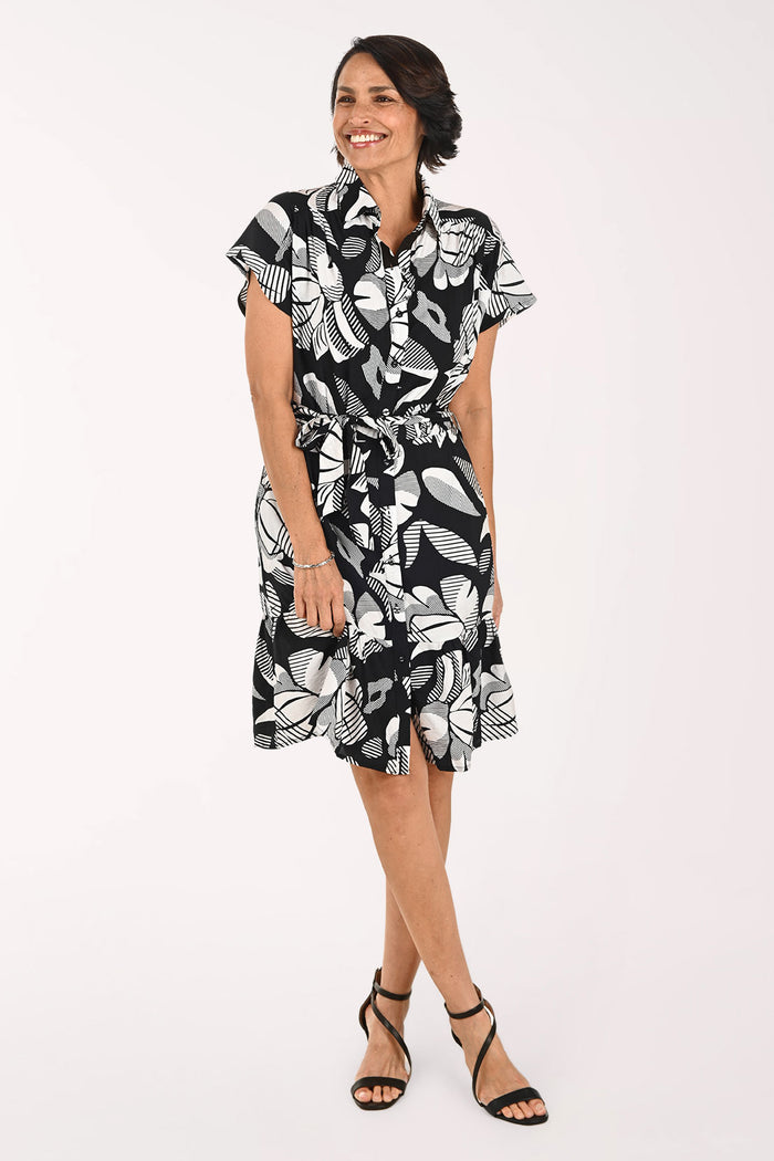 A woman modeling a short-sleeve black and white tropical floral print dress with a tie waist, paired with black strappy high-heeled sandals, against a plain white studio background.