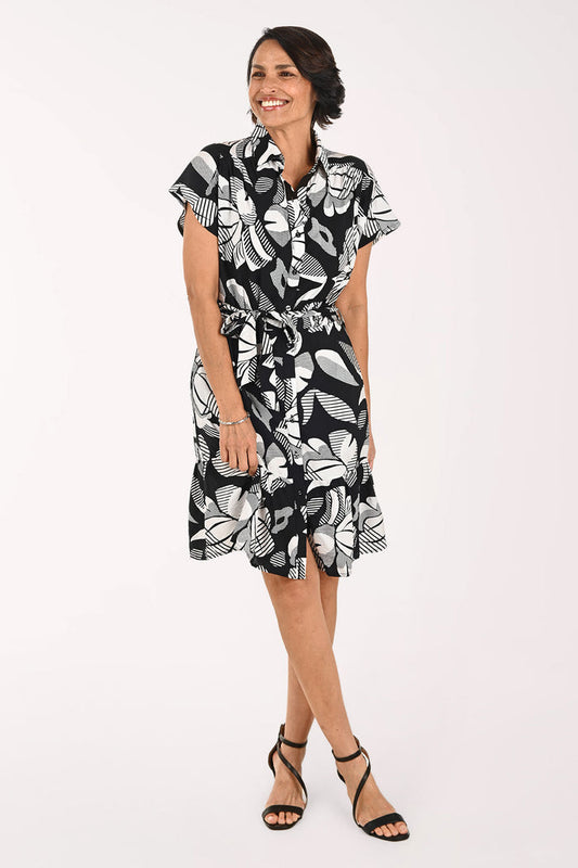 A woman modeling a short-sleeve black and white tropical floral print dress with a tie waist, paired with black strappy high-heeled sandals, against a plain white studio background.
