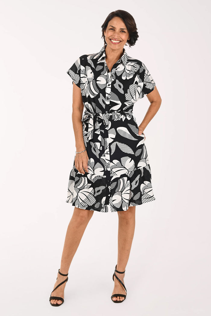 A woman modeling a short-sleeve black and white tropical floral print dress with a tie waist, paired with black strappy high-heeled sandals, against a plain white studio background.